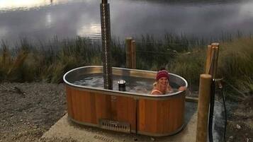 A hot tub with a metal pole and a wooden structure, situated on a gravel area near a body of water.