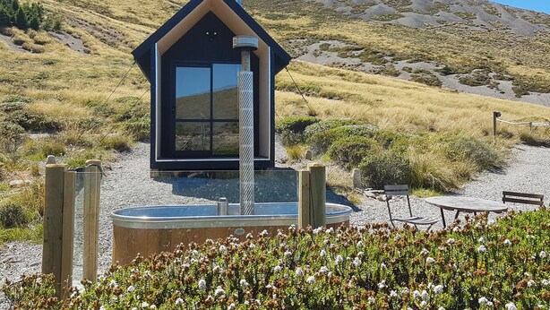 Una cabaña moderna de color oscuro con un gran ventanal, rodeada por un área de grava con una piscina pequeña y mobiliario exterior.