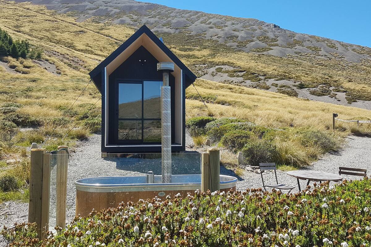 A modern, dark-colored cabin with a large glass door, surrounded by a gravel area with a small pool and outdoor seating.