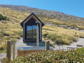Una cabaña moderna de color oscuro con un gran ventanal, rodeada por un área de grava con una piscina pequeña y mobiliario exterior.
