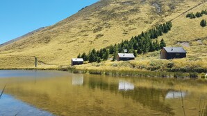 Una escena tranquila junto al lago con tres cabañas, agua cristalina que refleja los alrededores y una montaña al fondo.