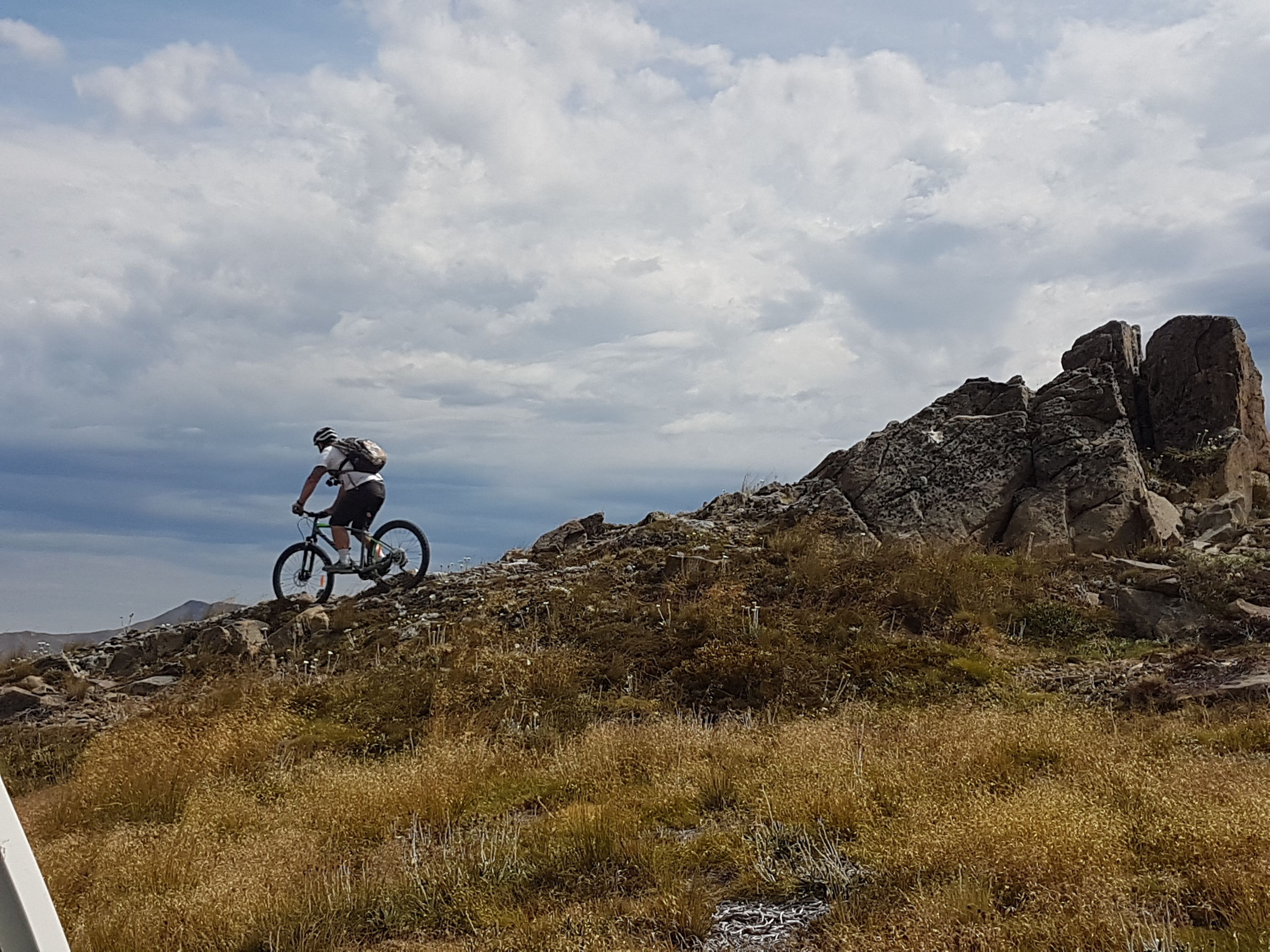 Un ciclista de montaña en un sendero rocoso, con vegetación escasa y una gran formación rocosa al fondo.