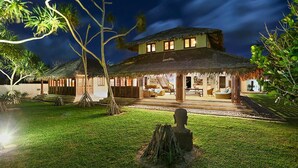 A two-story house with a thatched roof, a staircase leading to the upper level, and a well-maintained lawn.