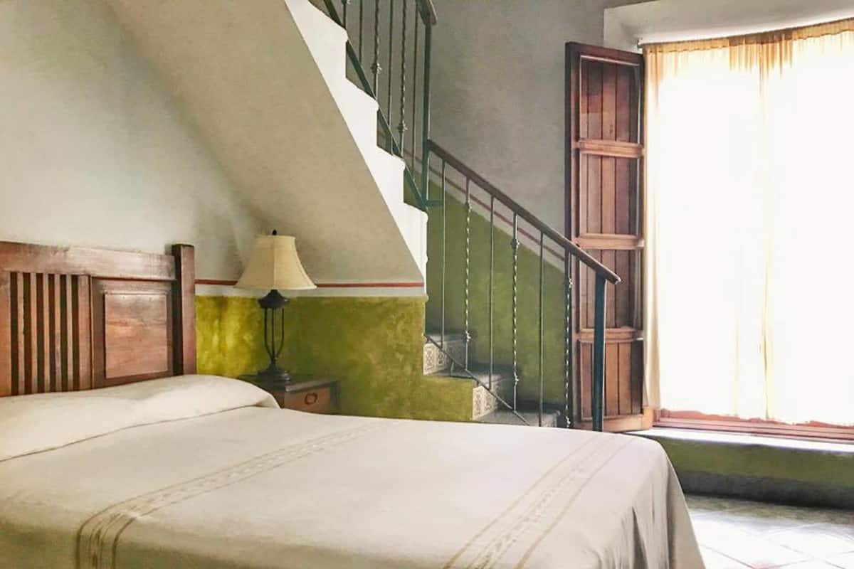 Un dormitorio con una cama, una mesita de noche, una lámpara, una escalera y un ventanal con cortinas.