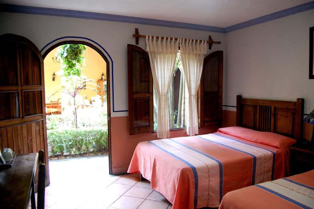 Habitación con dos camas, muebles de madera y vistas a un espacio exterior con plantas a través de un arco abierto.