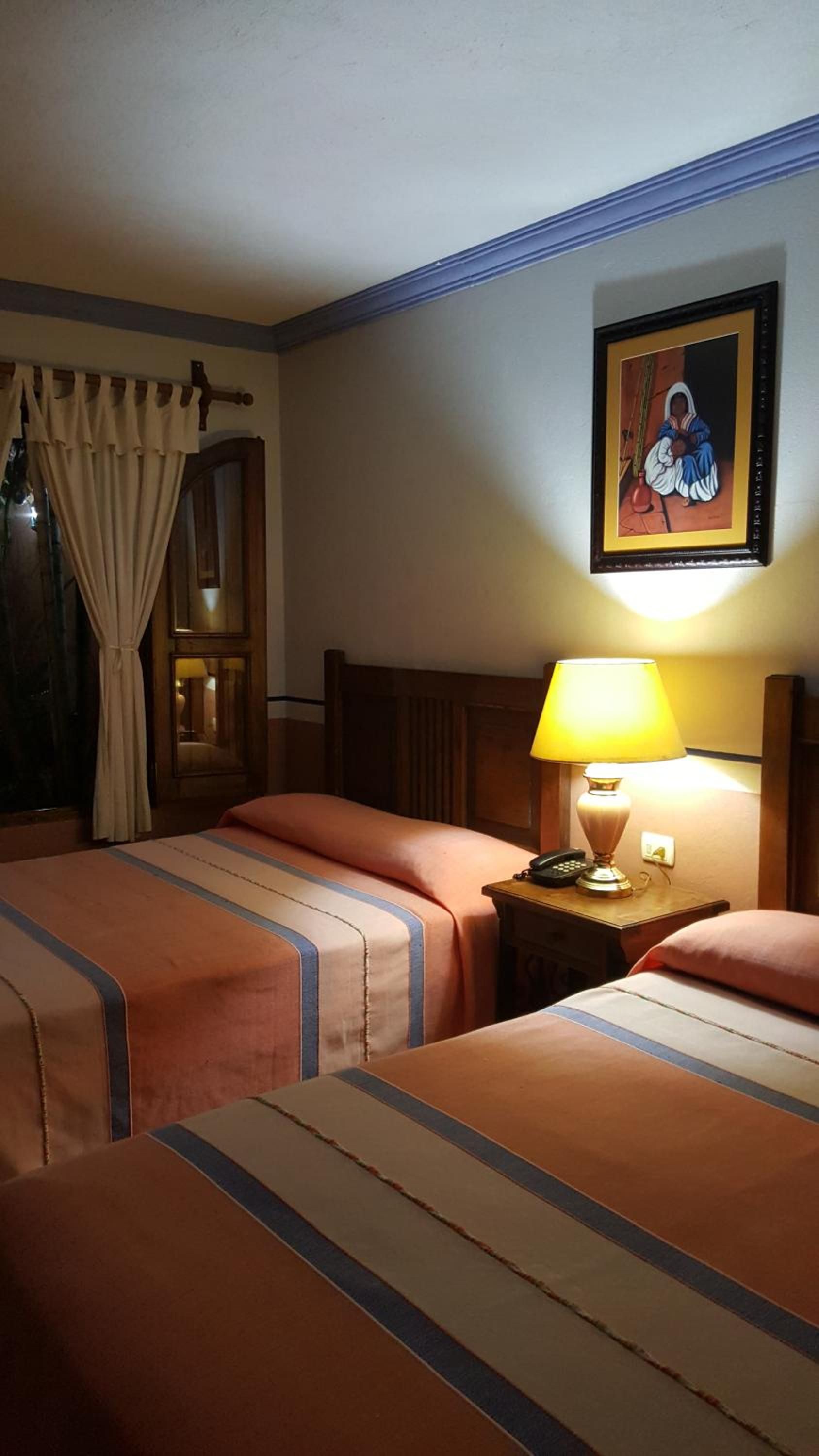 Habitación de hotel con dos camas, una mesita de noche con lámpara, un cuadro enmarcado en la pared y una ventana con cortinas.