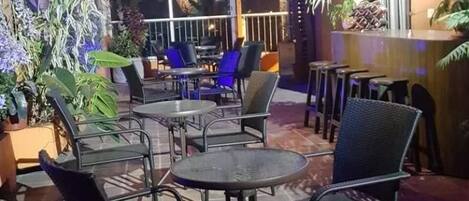 Zona salotto all'aperto con tavoli e sedie, circondata da piante in vaso e verdeggianti decorazioni pensili.