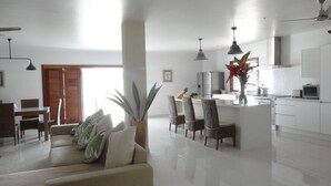 A modern living room with a sofa, chairs, and a dining area. There is a kitchen with white cabinets and a dining table. A large window allows natural light to fill the space. - Paradise Point Views (Port Vila)