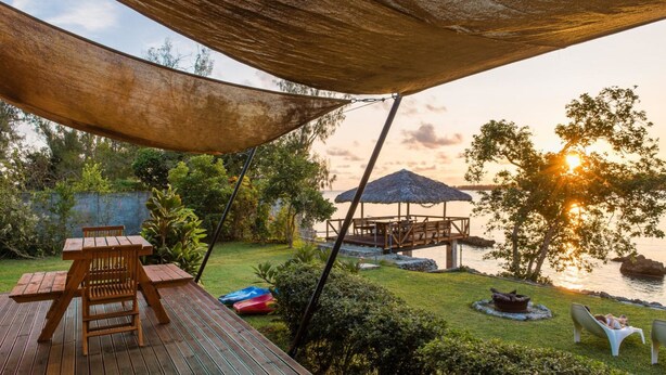 A wooden deck with a table and chairs, a thatched umbrella, and a view of the sunset over water. - Teouma Beachfront (Teouma)