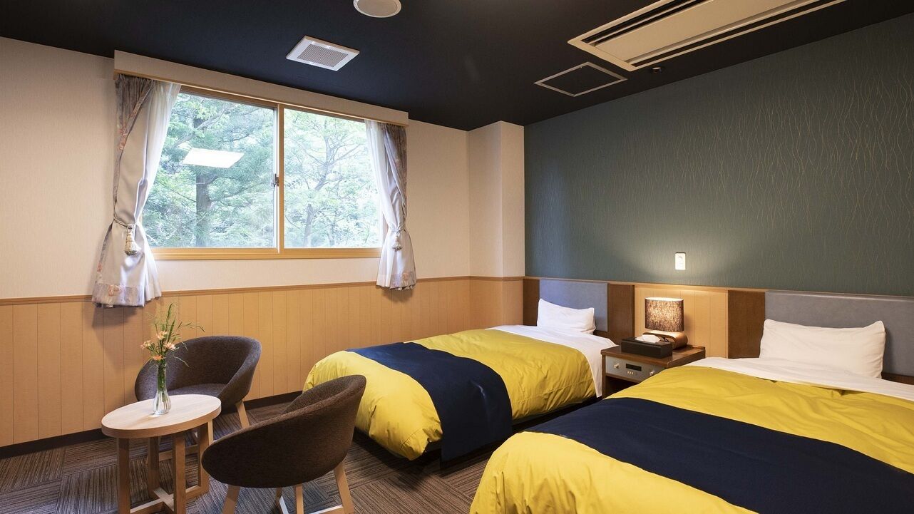 Een hotelkamer met twee bedden, een tafeltje, een stoel en een raam met gordijnen.