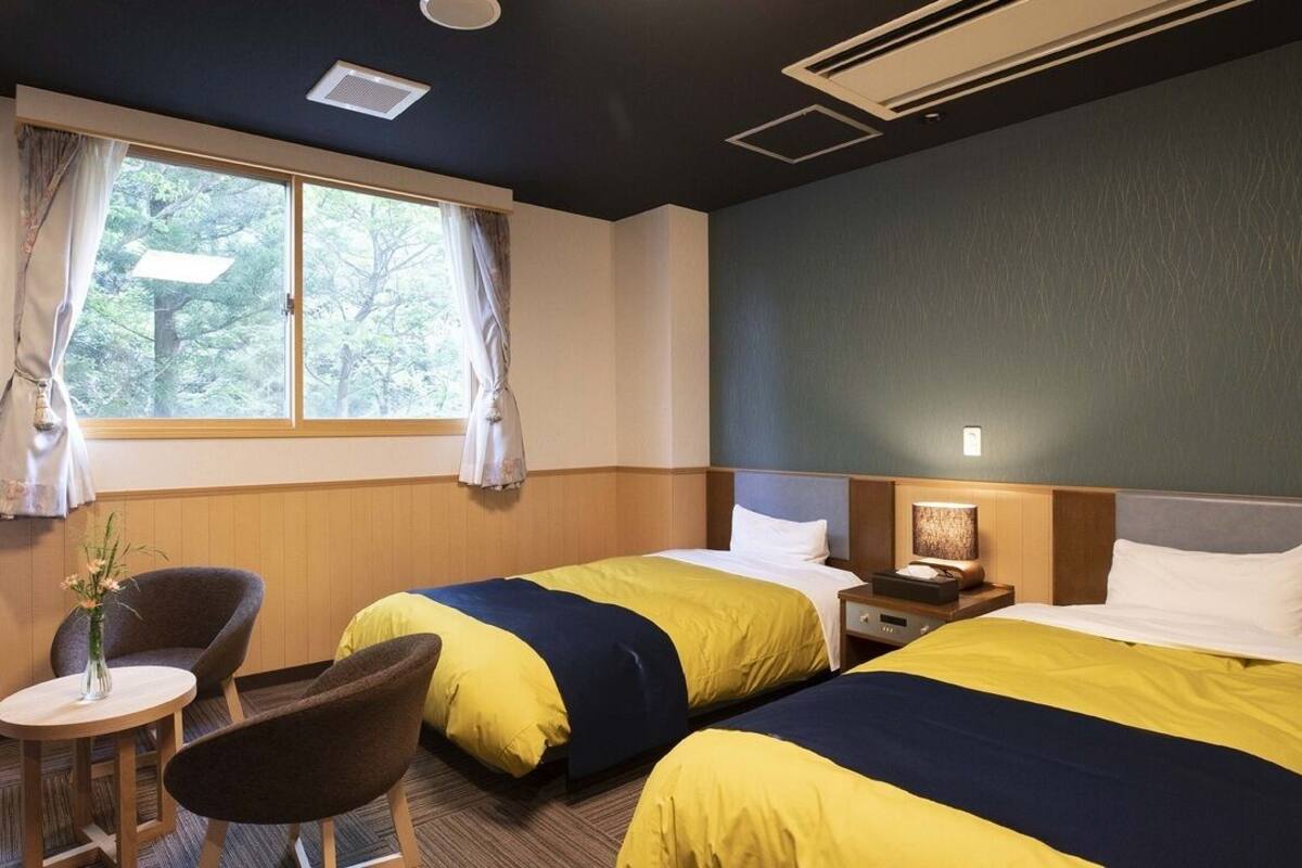 A hotel room with two beds, a small table, a chair, and a window with curtains.