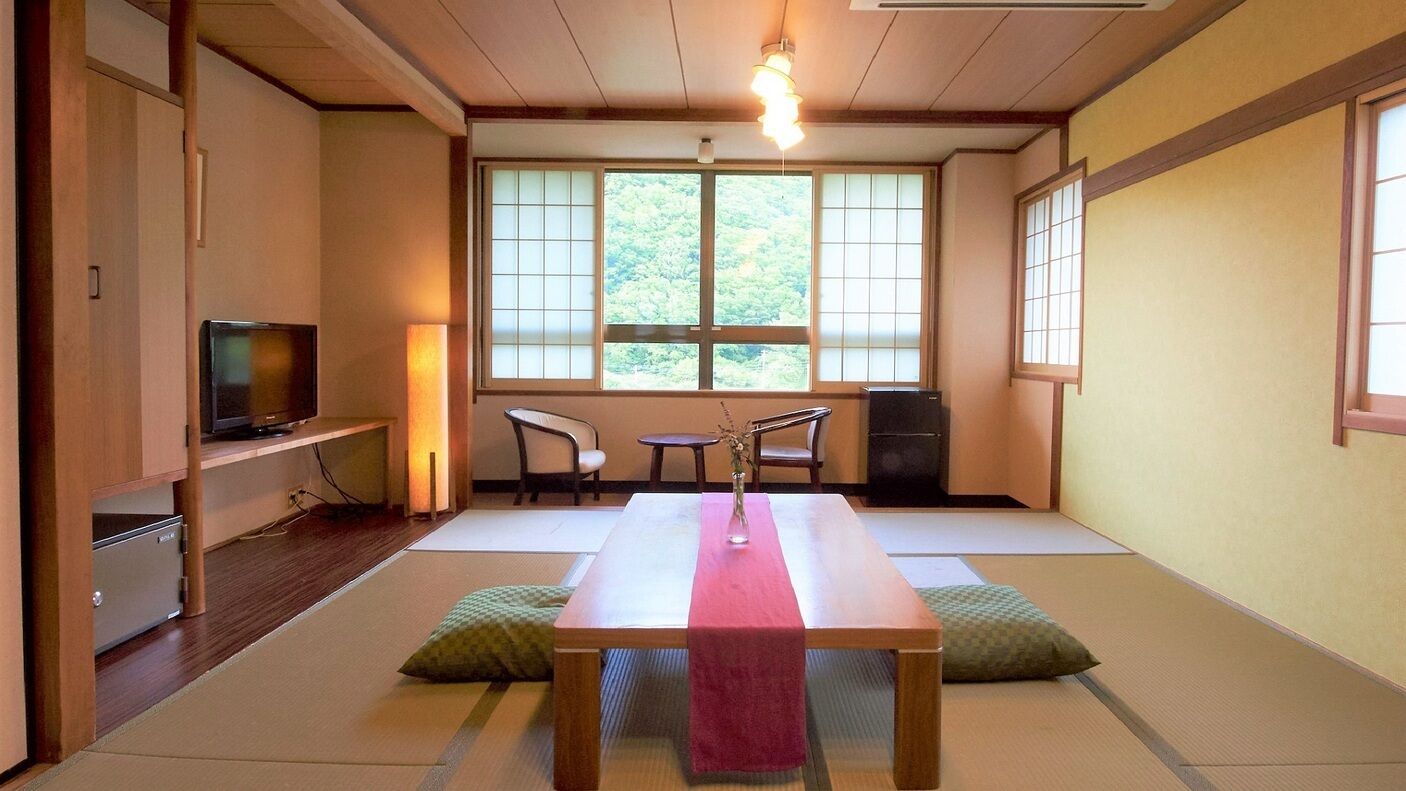 Een traditionele Japanse kamer met tatami vloeren, een houten tafel, een televisie en uitzicht op een bos door grote ramen.
