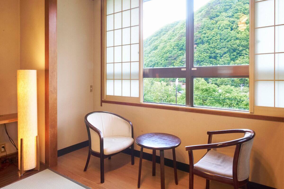 A room with a view of a lush green hill through a large window with wooden frames and glass panes. There are two chairs and a small round table in the room.