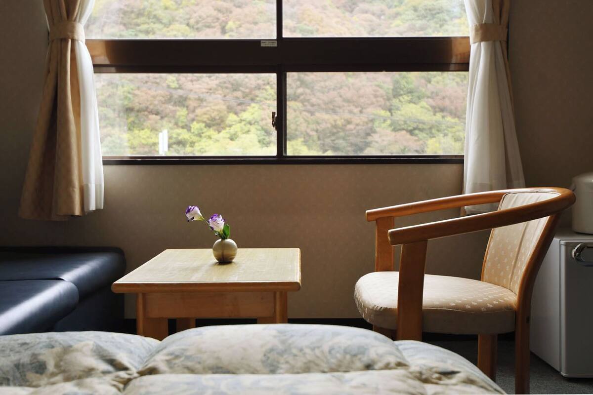 A room with a bed, a wooden chair, a small table with a vase of flowers, and a window with curtains.