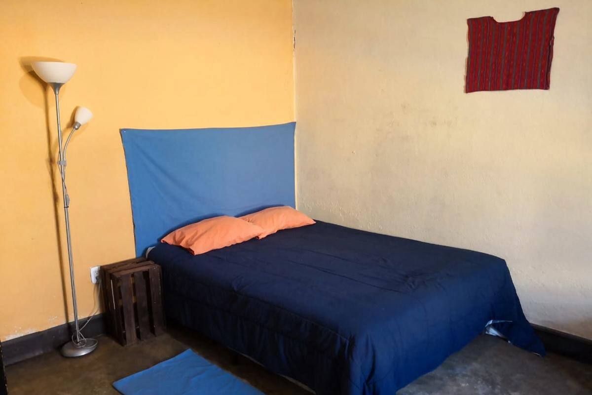 A single bed with a blue and orange bedspread, a small bedside table, a floor lamp, and a red curtain on the wall.