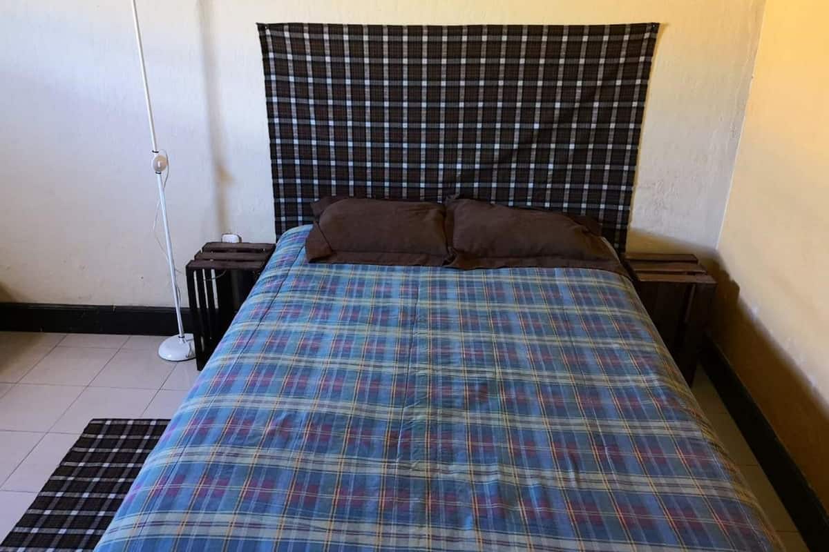 A bedroom with a plaid bedspread, two pillows, a bedside table, and a floor lamp.