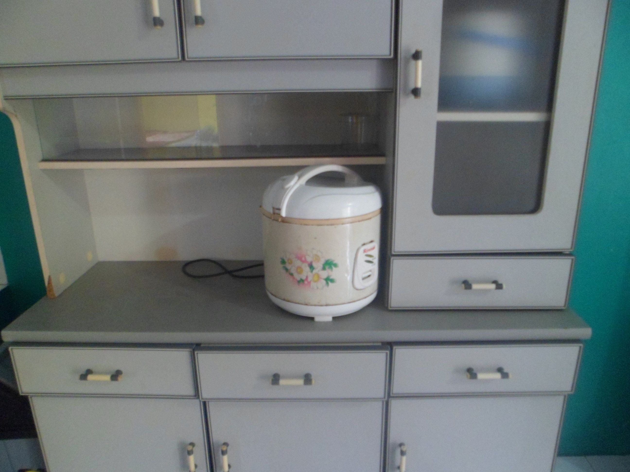 Una cocina con un hervidor eléctrico blanco sobre una encimera gris, un microondas encima y un armario gris con puerta de cristal.
