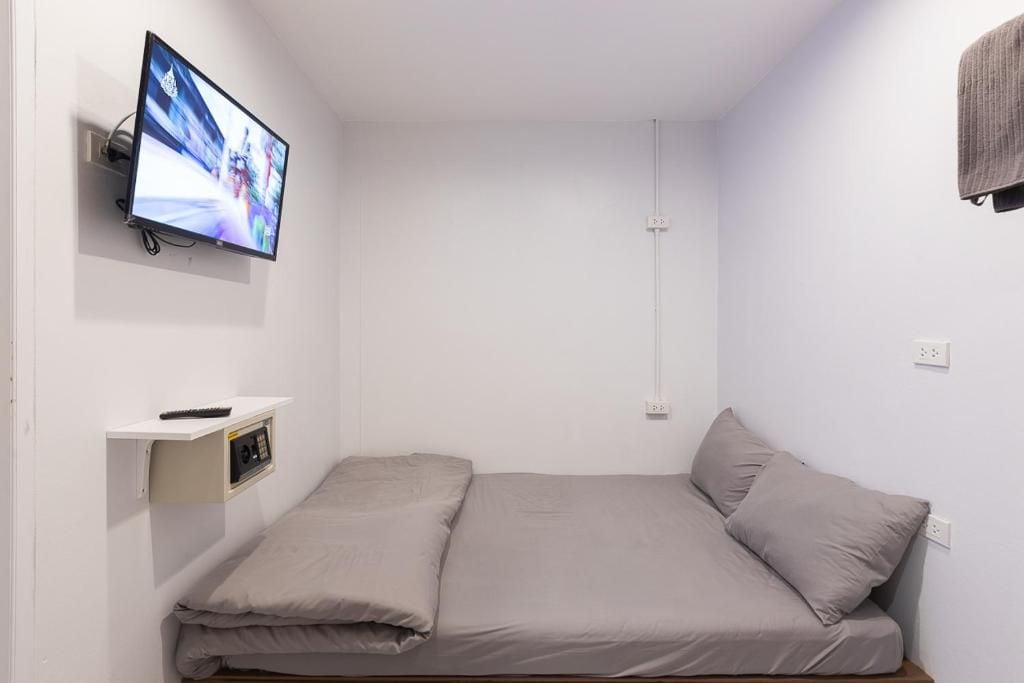 Una habitación pequeña con una cama individual, un televisor montado en la pared, un enchufe y un estante con el control remoto.