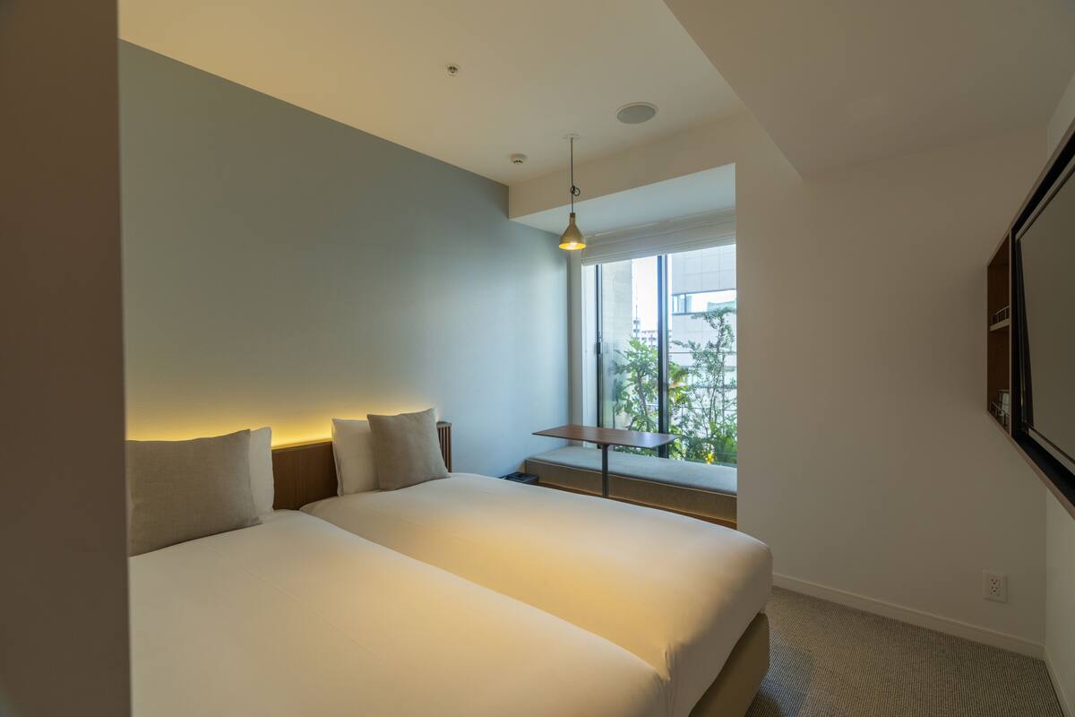 Una habitación de hotel moderna con una cama grande, una mesita pequeña y un ventanal con vista a espacios verdes.