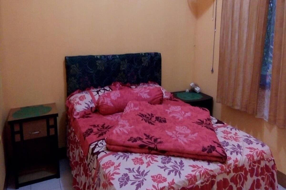 Une chambre à coucher avec un lit, une table de chevet, une horloge murale, un tableau encadré et une fenêtre avec des rideaux.