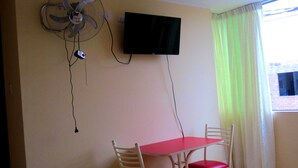 A room with a TV mounted on the wall, a table with two chairs, a standing fan, and a window with curtains. - Hostal Sandel (Barranca)