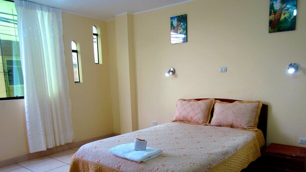 A neatly made bed with a white duvet and two pillows, a small bedside table with a lamp, and a window with curtains. - Hostal Sandel (Barranca)