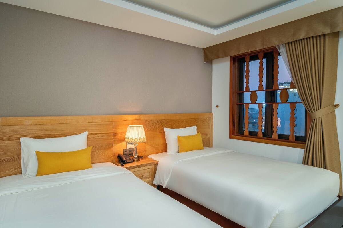 A hotel room with two beds, a wooden headboard, a nightstand with a lamp, and a window with curtains.