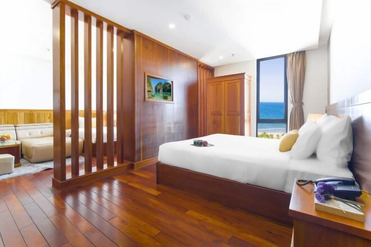 A hotel room with a wooden floor, a bed with white bedding, a nightstand with a lamp, and a large window with a view of the sea.