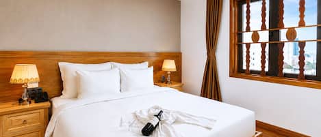 Habitación de hotel con una cama grande, dos mesitas de noche con lámparas, un cabecero de madera, un ventanal con vistas y una cama bien hecha con bata y botella de vino.