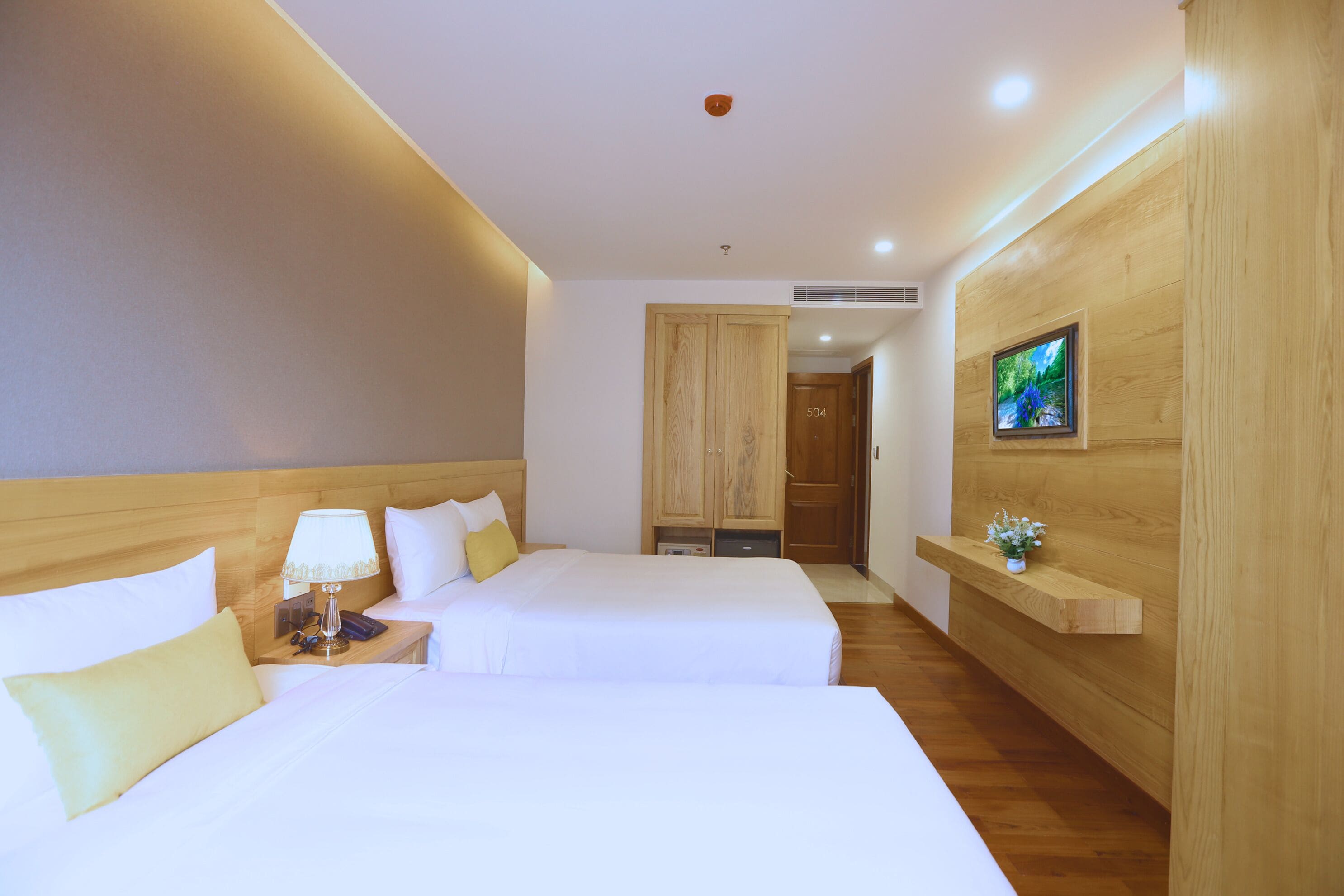 A hotel room with two beds, a nightstand with a lamp, a wooden wardrobe, a TV on the wall, and a small plant on a shelf.