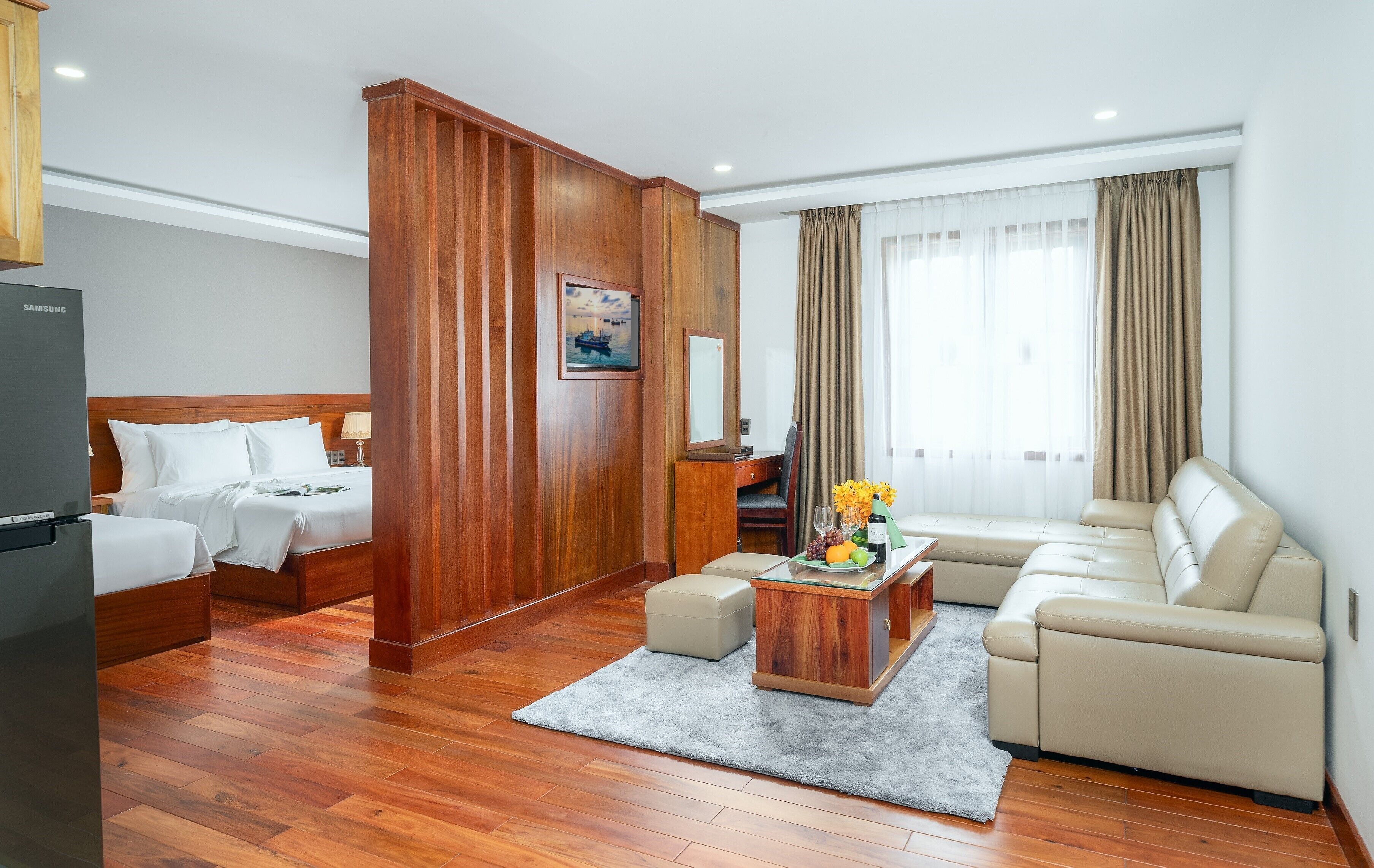 Une chambre d’hôtel avec un plancher en bois, un lit, un canapé, une table basse avec des fruits et une grande fenêtre avec des rideaux.