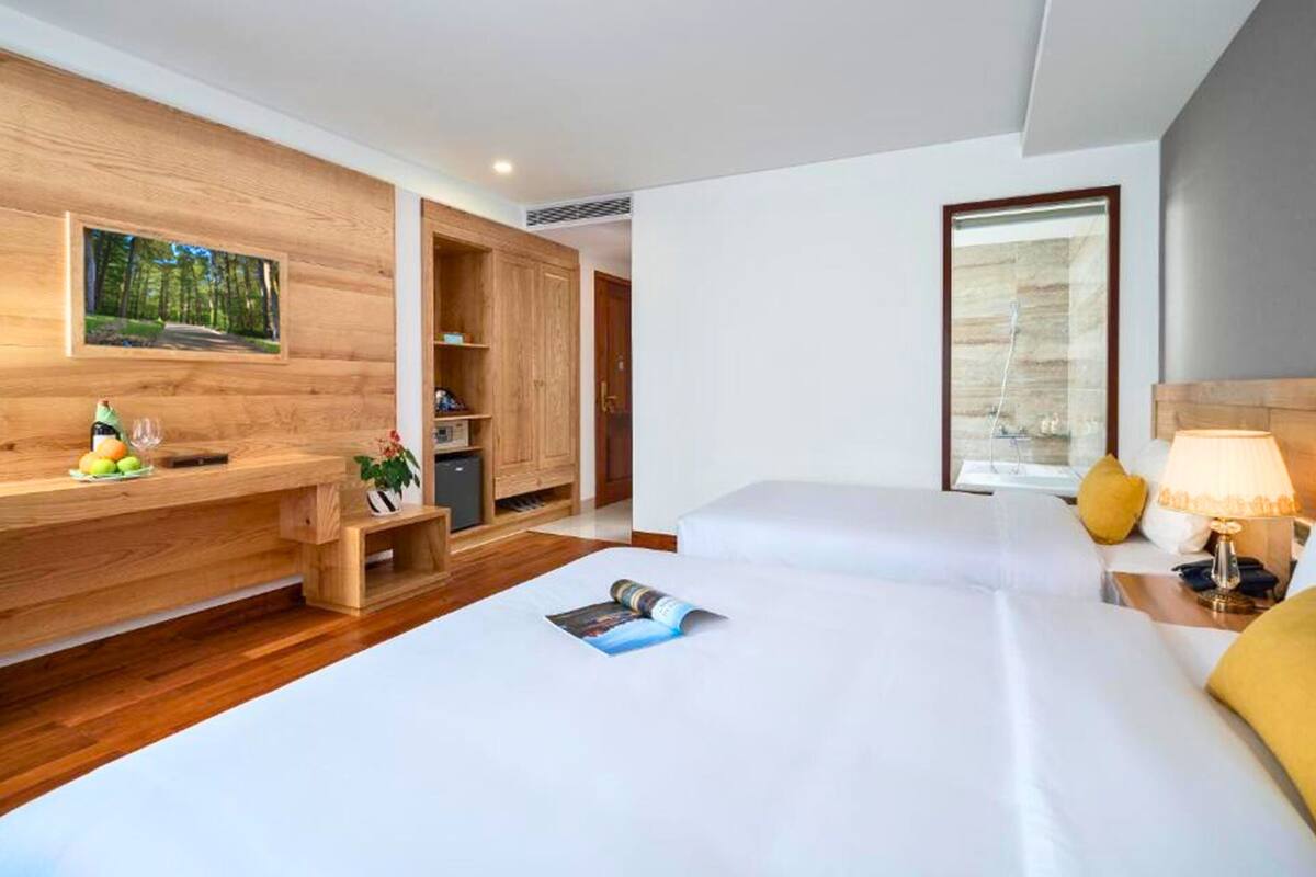 A hotel room with a wooden desk, a bed with white linens, a bedside table with a lamp, and a window with a view of a building.
