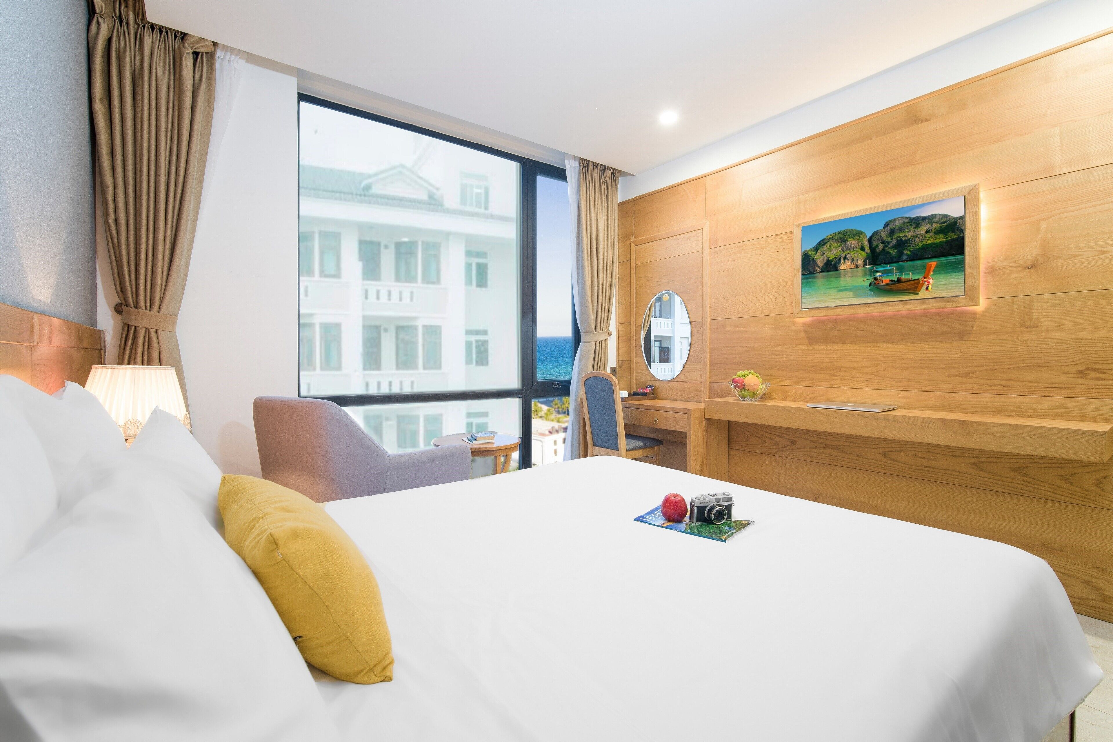 A hotel room with a large bed, a desk, a chair, a round mirror, and a view of the sea through a large window.
