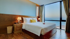 A hotel room with a large bed, wooden headboard, bedside tables with lamps, a wicker basket, and a view of the sea through a large window.
