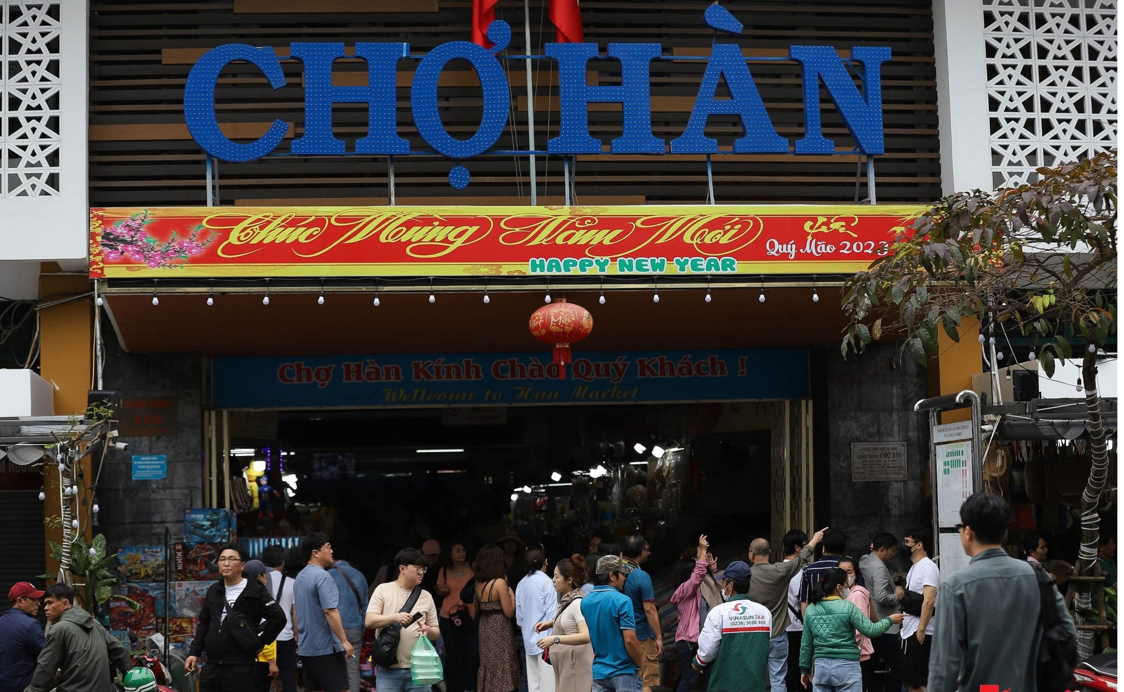A busy entrance to a market with a prominent sign reading "CHQHAN" and a festive banner for the New Year.