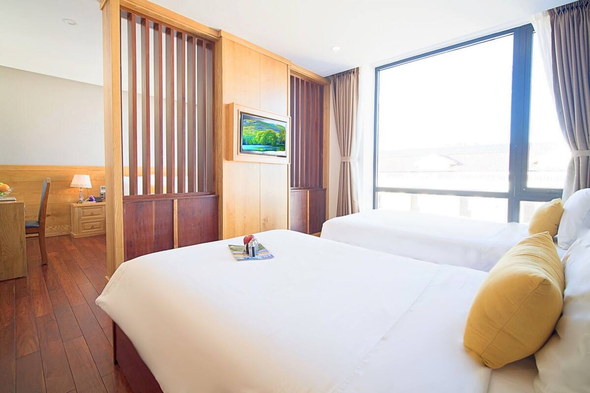 A hotel room with a large bed, a desk with a lamp, a TV on the wall, and a window with curtains.