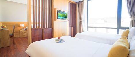 A hotel room with a large bed, a desk with a lamp, a TV on the wall, and a window with curtains.