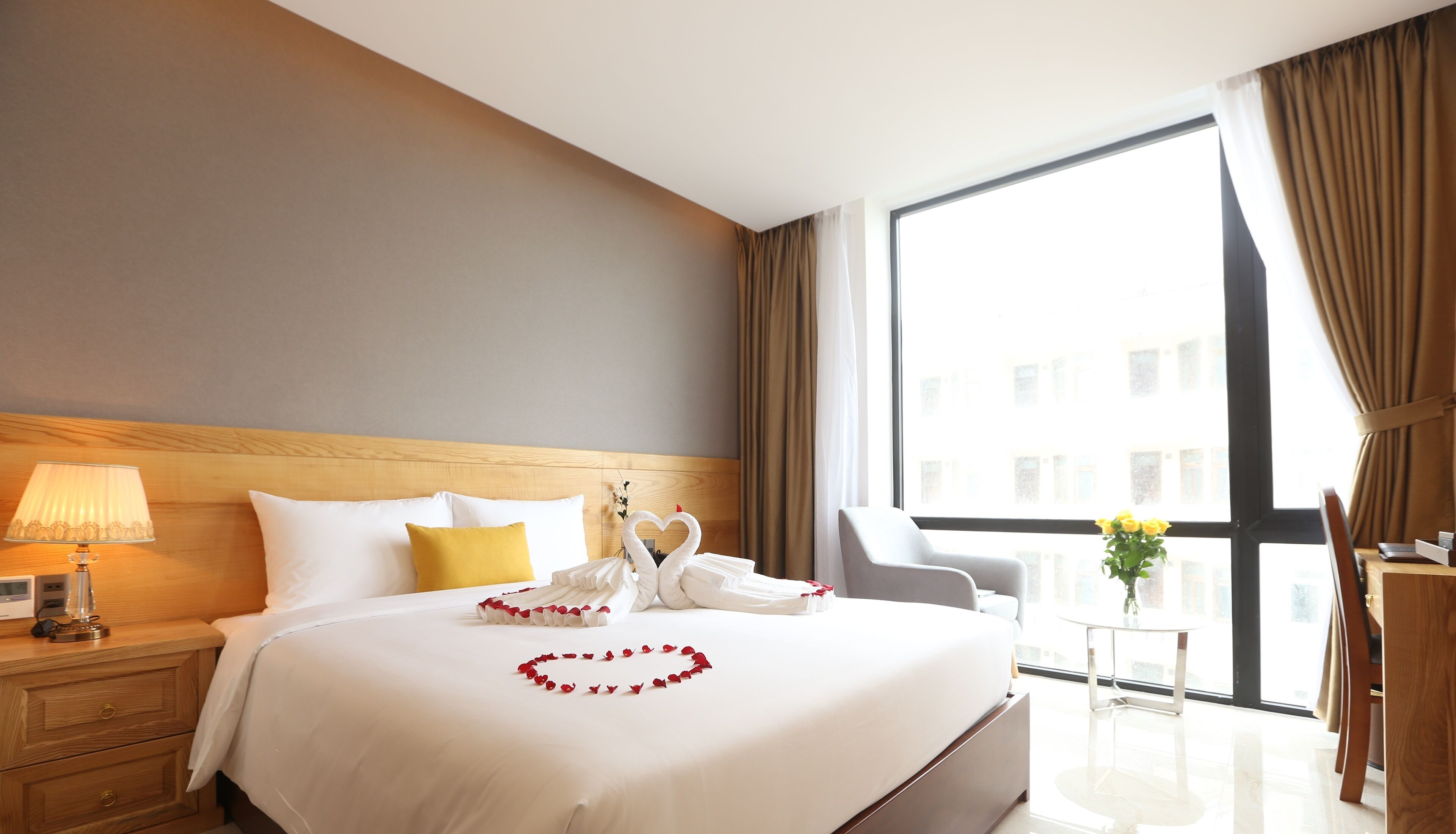 A hotel room with a large bed, a nightstand with a lamp, a chair, a small table with a vase, and a view of buildings through a large window.