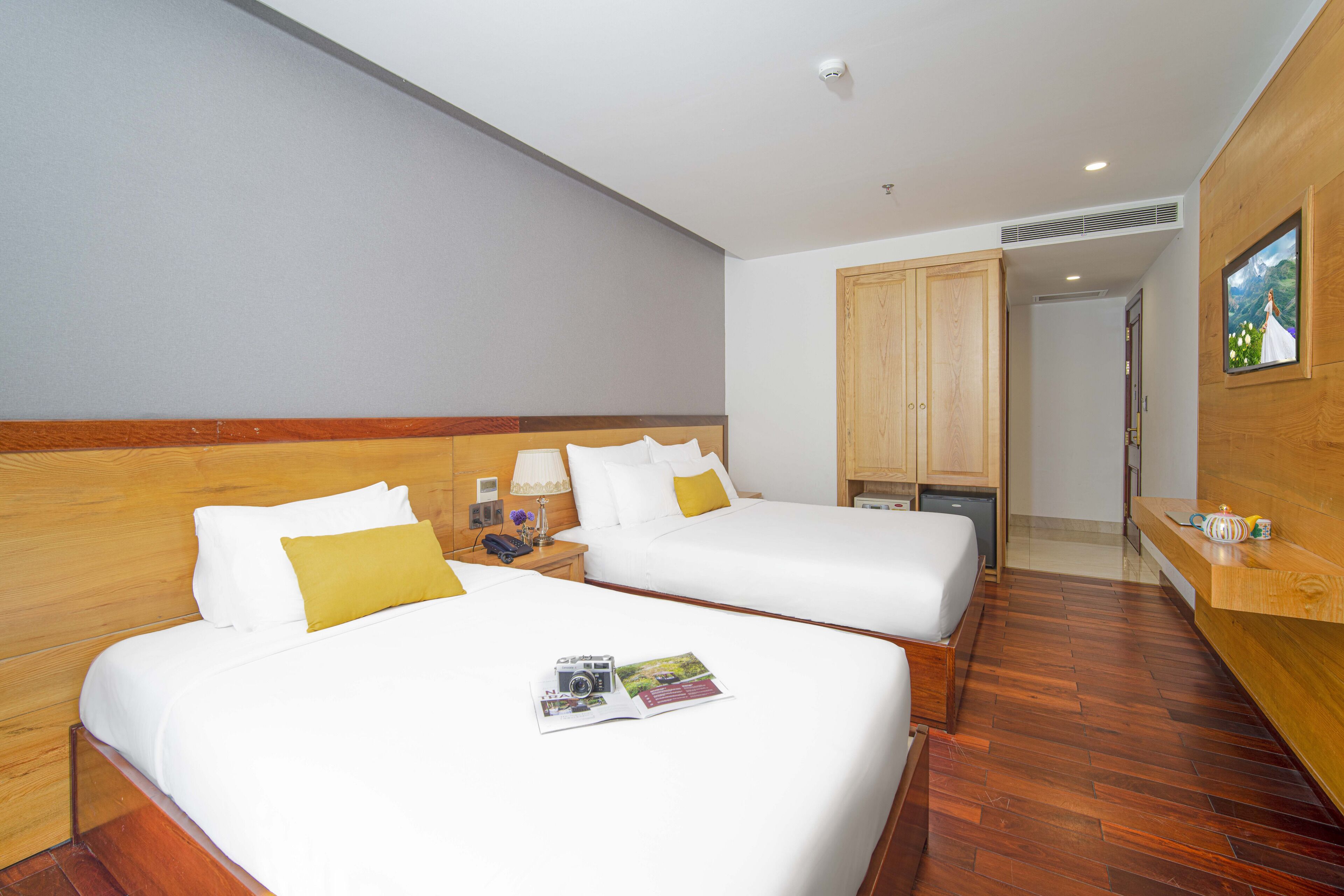 Een hotelkamer met twee bedden, houten hoofdeinden, een houten vloer, een houten kledingkast, een tafeltje met een lamp, en een lijst met een afbeelding aan de muur.