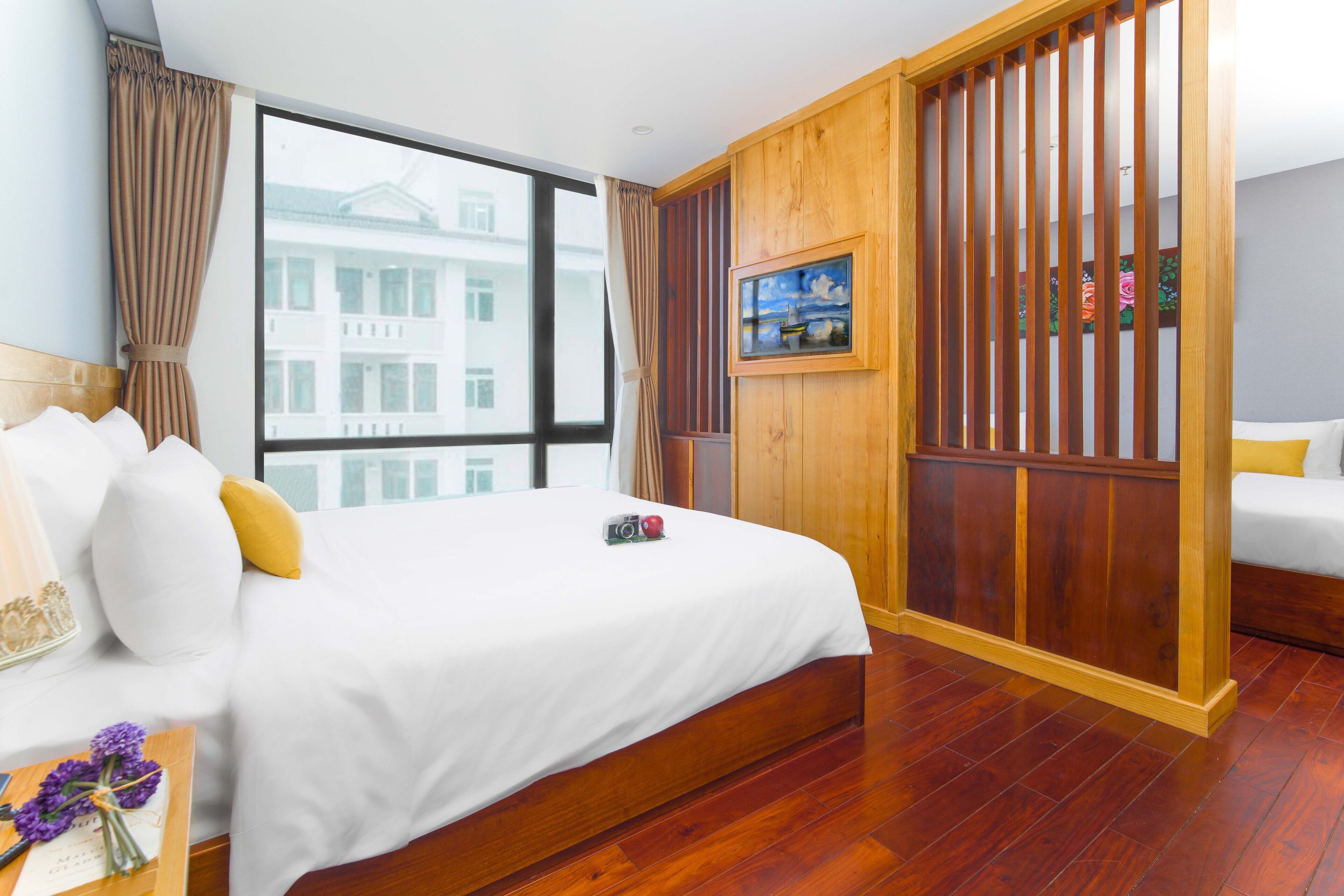 Habitación de hotel con una cama grande, un separador de madera, una ventana con vistas a un edificio y un cuadro en la pared.