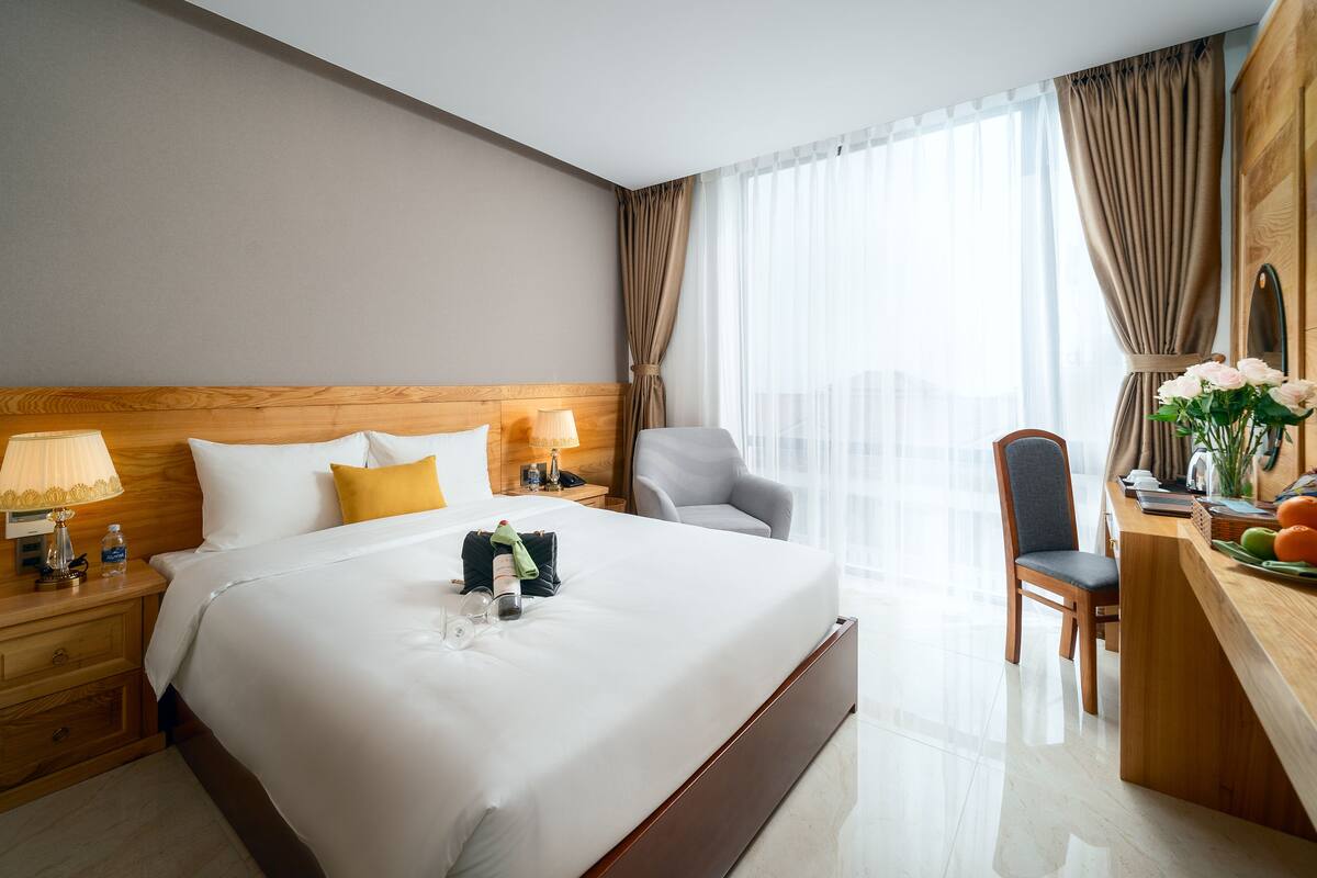 A hotel room with a large bed, wooden headboard, two bedside lamps, a desk with a chair, and a view of the city through sheer curtains.