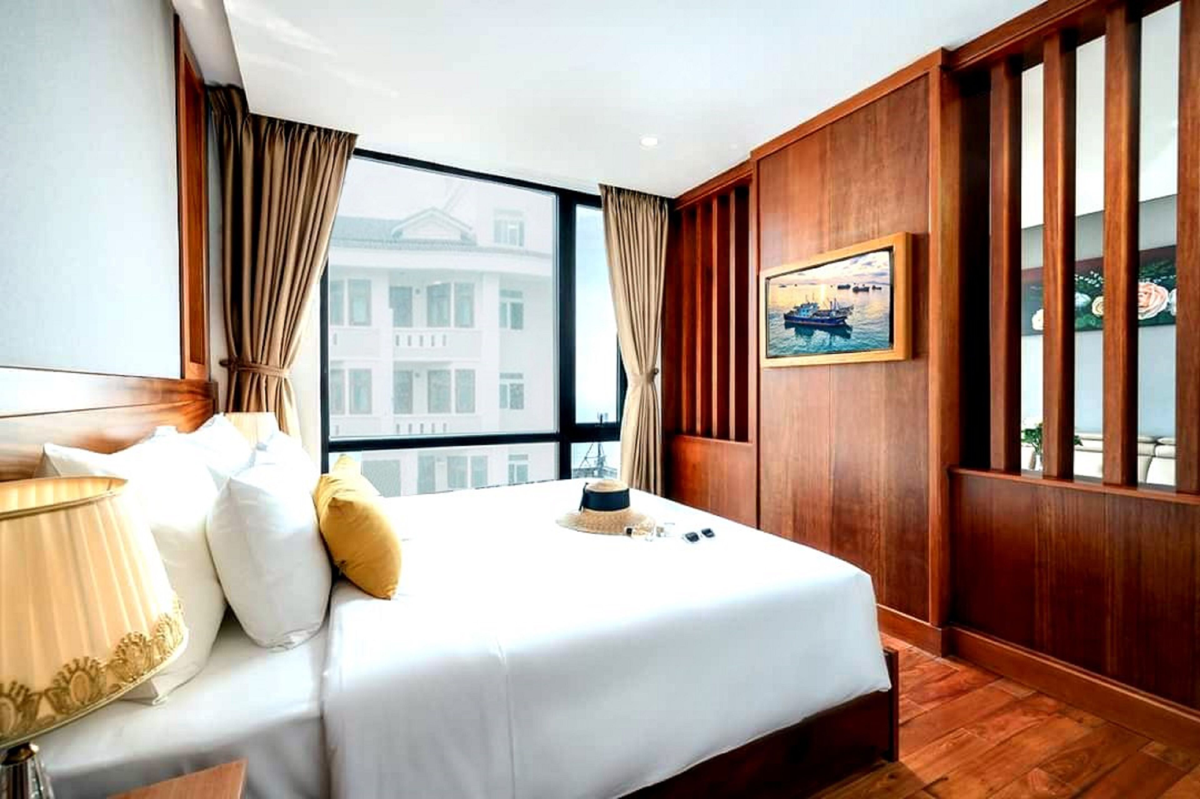 A hotel room with a large bed, wooden paneling, a window with a view of buildings, and a painting on the wall.