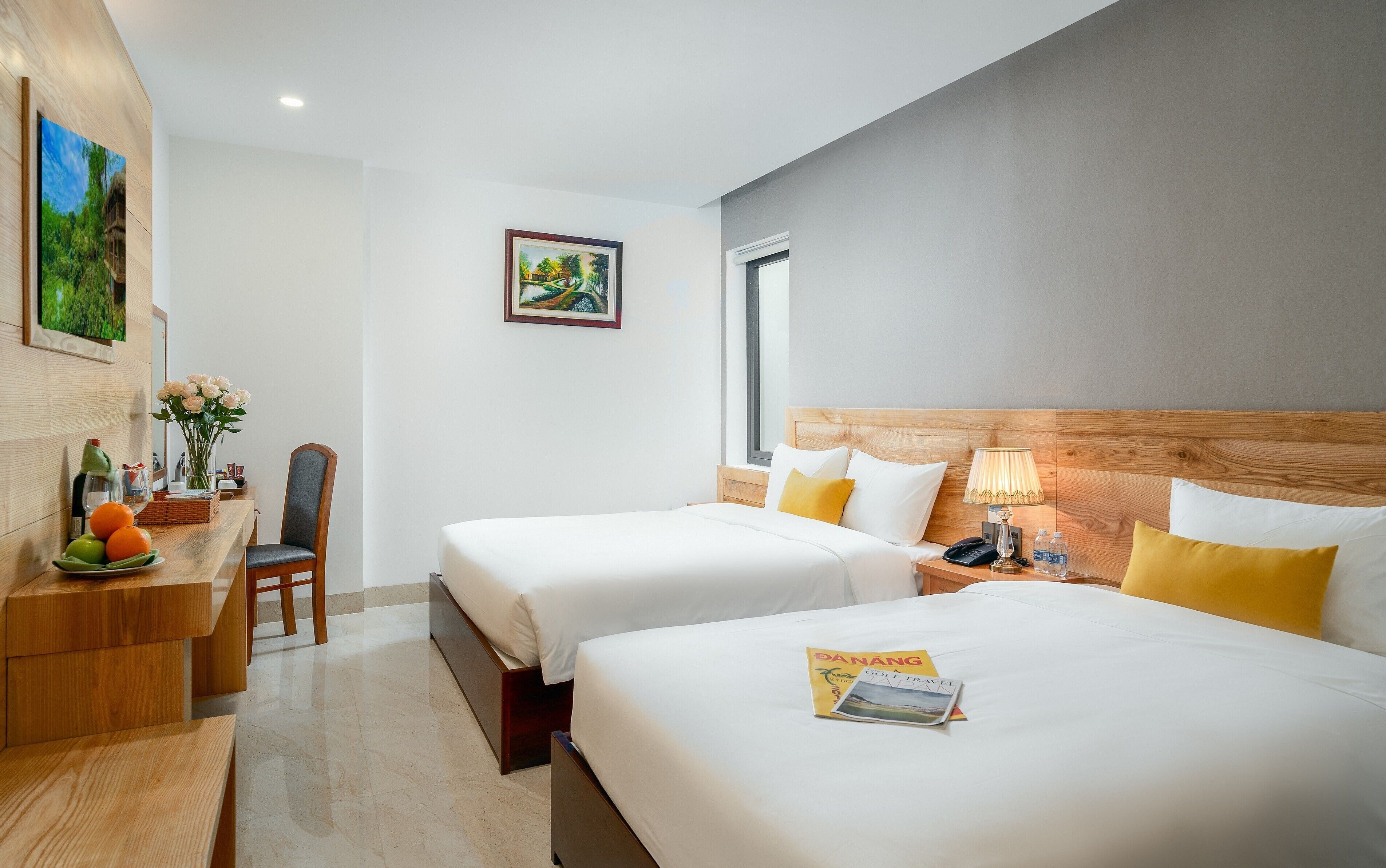 A hotel room with two beds, a desk with a chair, a vase of flowers, and a painting on the wall.