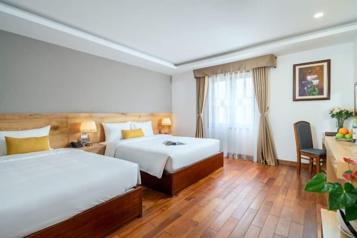 A hotel room with two beds, wooden flooring, a desk with a chair, and a window with curtains.