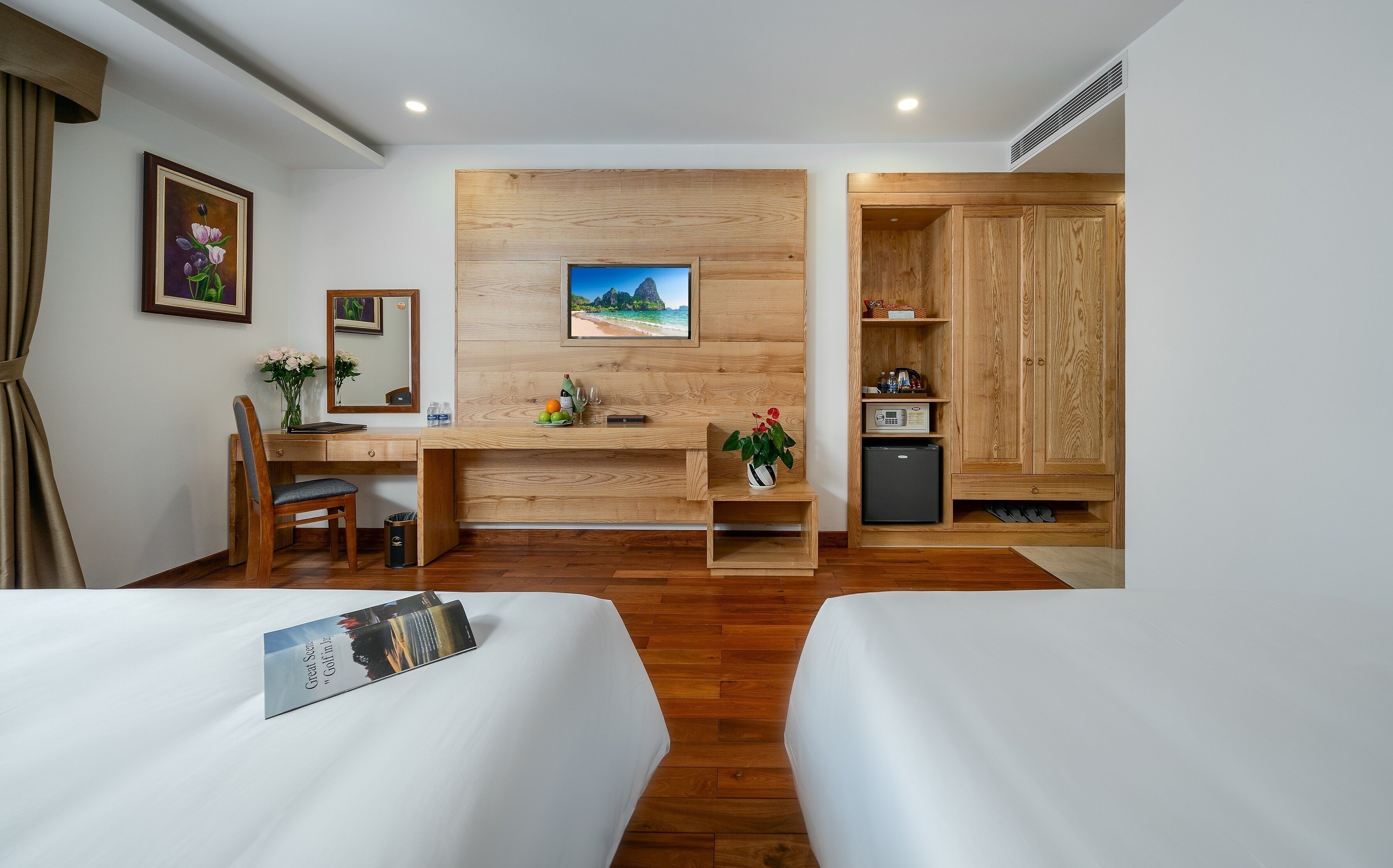 A hotel room with two beds, a wooden desk, a small refrigerator, a mirror, and a painting on the wall.