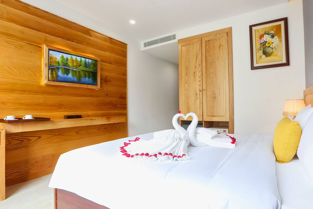 A hotel room with a wooden wall, a bed with white bedding, a heart-shaped towel arrangement, a painting, and a yellow lamp.