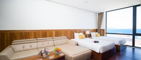 A modern hotel room with a sofa, a bed, a small table with fruit and flowers, and a large window offering a view of the sea.