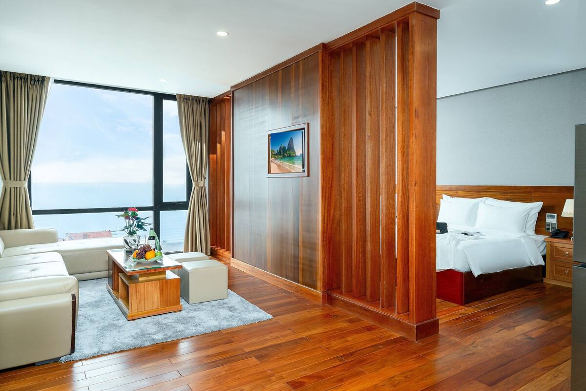 A modern hotel room with a large window, wooden paneling, a bed, a sofa, a coffee table, and a small table with fruit.