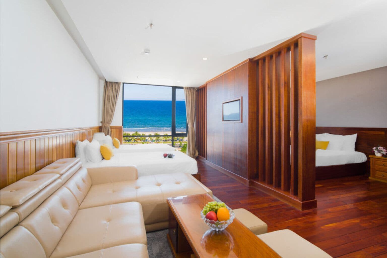 A hotel room with a large bed, a sofa, a dining table with fruit, and a view of the beach.