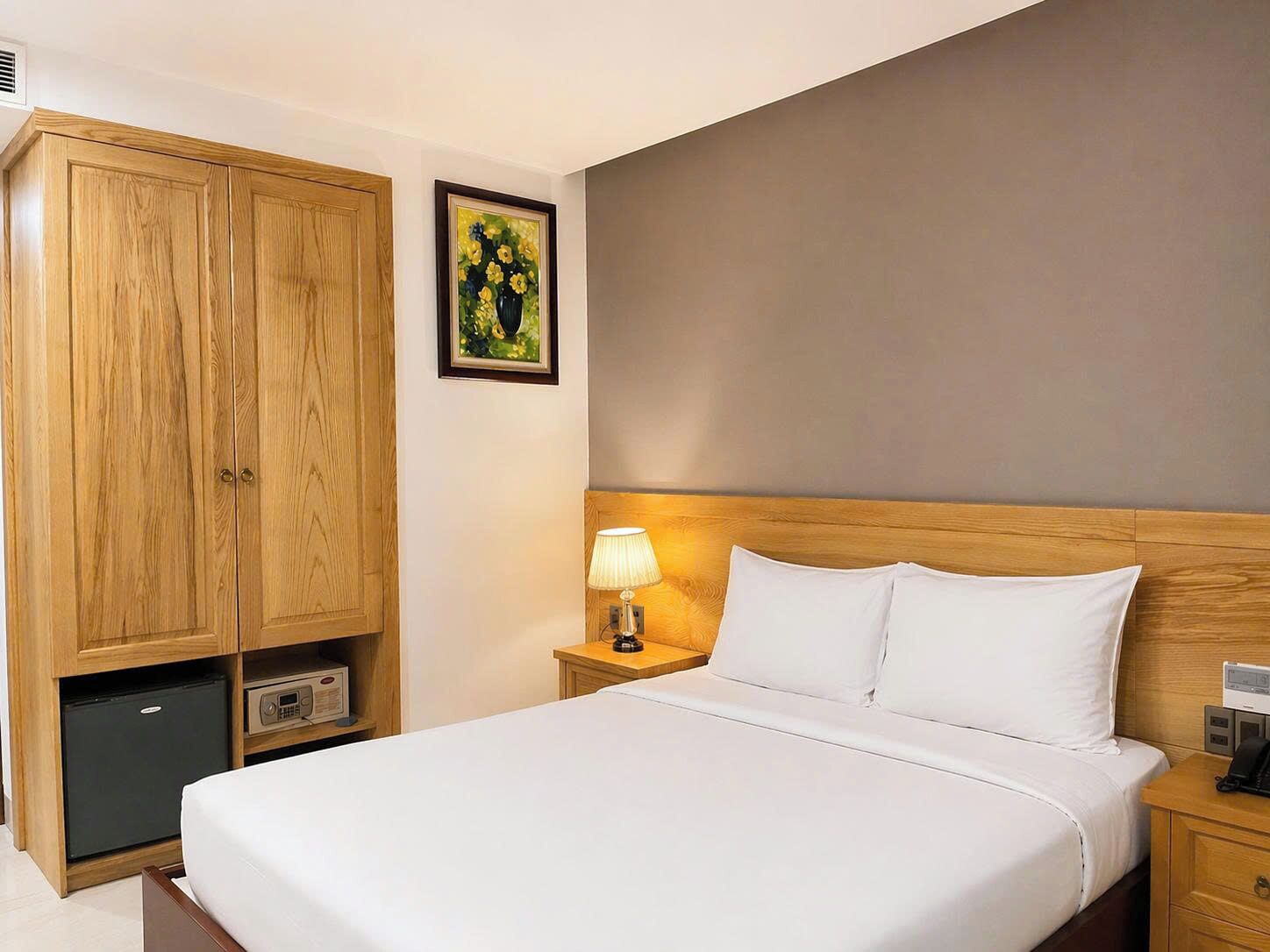 Una habitación de hotel con una cama grande, un armario de madera, una mesita de noche con una lámpara y un cuadro en la pared.
